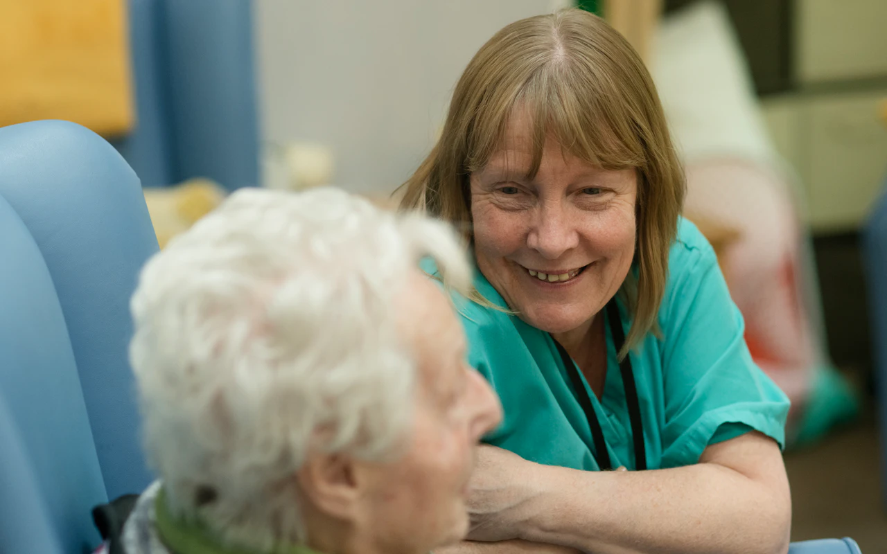 A nurse in green scrubs leaning in to talk with an older woman seated beside her.