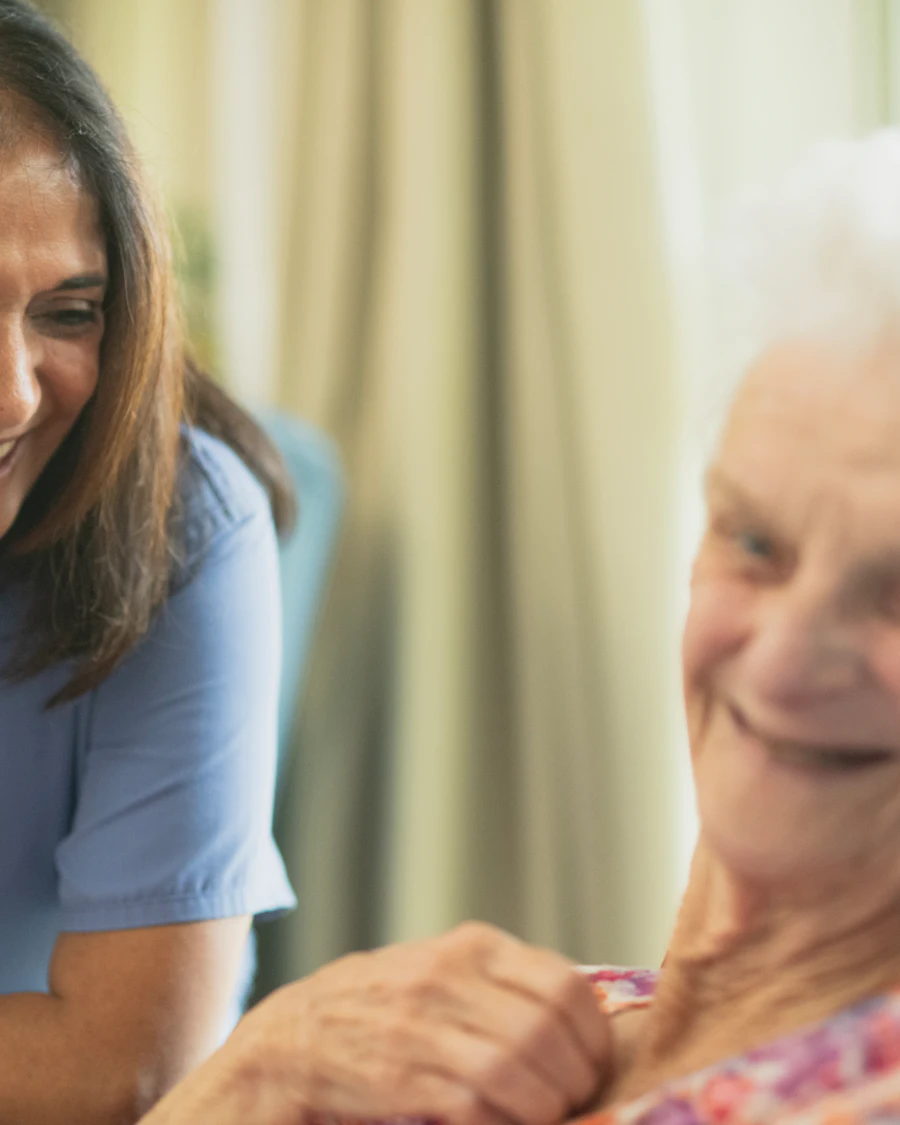 A care worker leaning close to an older woman, both of them looking down at something between them.