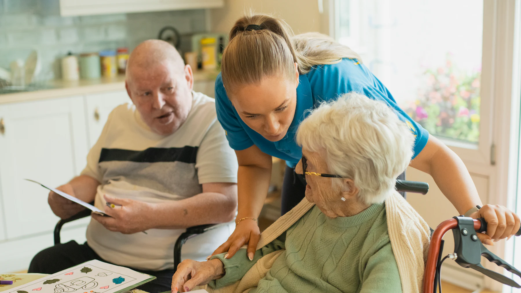A care worker leaning between an older man and an older woman at a table, the three of them working on a colouring sheet together.