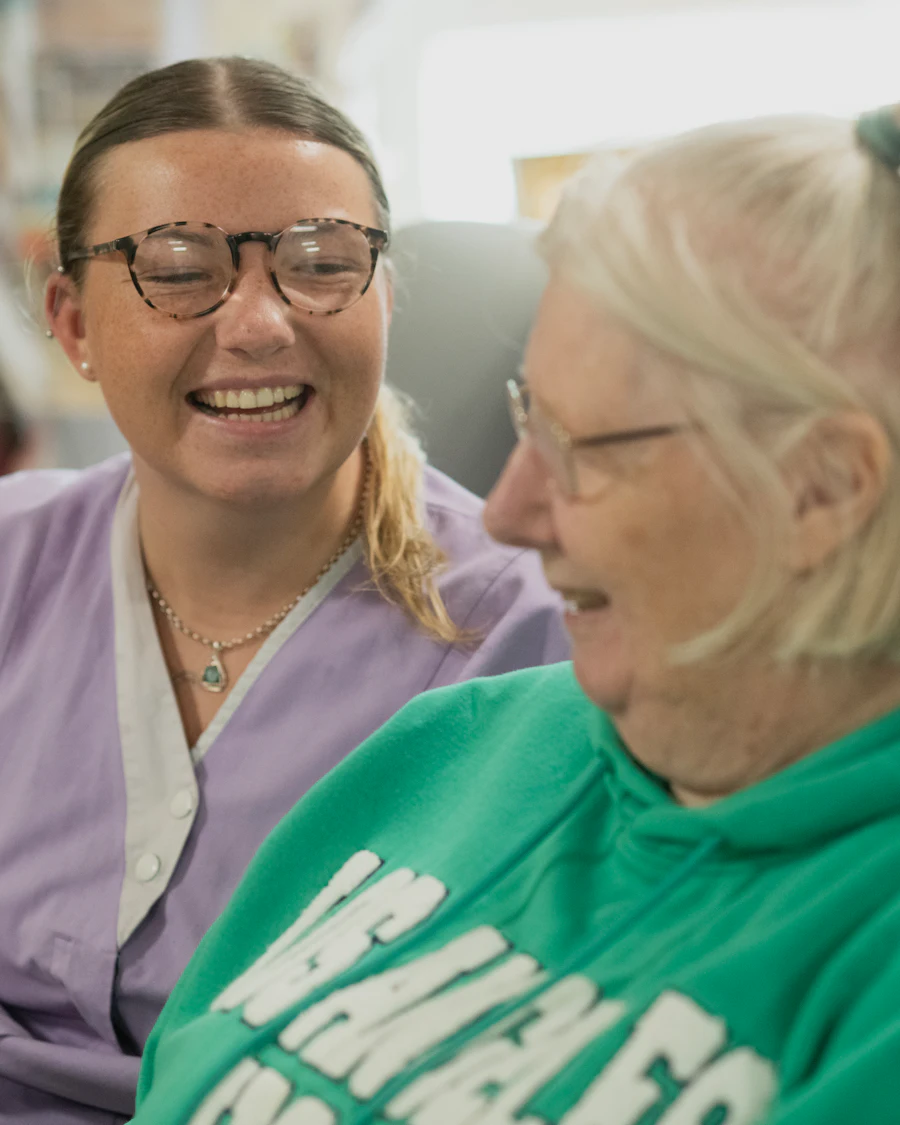 A care worker and an older woman laughing together, heads close, in a bright room.