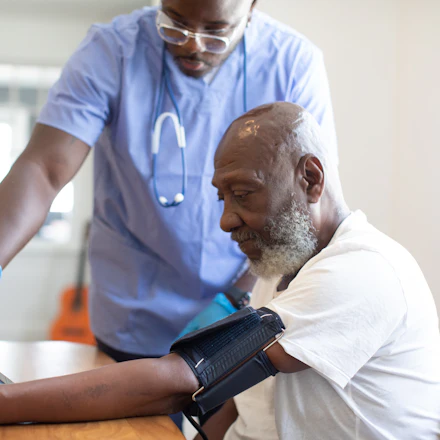A nurse in blue scrubs taking an older man's blood pressure in his own living room.