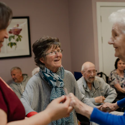 Older adults standing in a loose circle in a day room, holding hands with a member of staff among them.