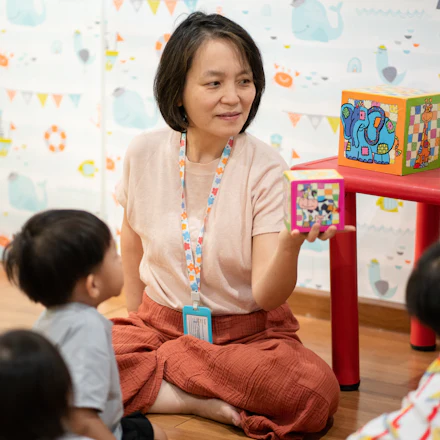An early childhood teacher sitting on the floor with a group of small children, holding up a stack of coloured blocks.
