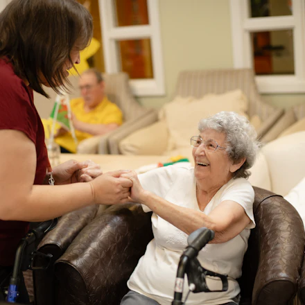 A care worker crouching to hold both hands of an older woman seated in an armchair, her wheeled walker parked in front of her.