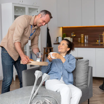 A support worker setting a hot drink down beside a woman sitting in a living room.