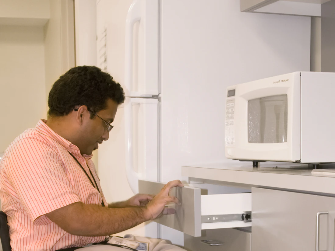 A man using a wheelchair reaching up to the microwave in his own kitchen.